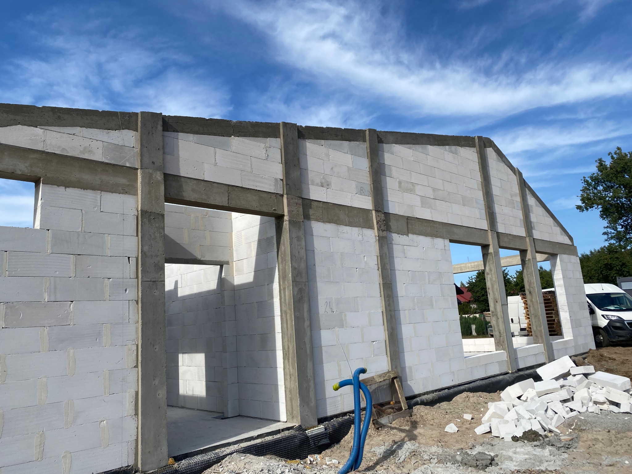 Surowy stan domu z betonu komórkowego, widoczne ściany, otwory okienne i drzwiowe, niebieskie rury na zewnątrz, w tle błękitne niebo.