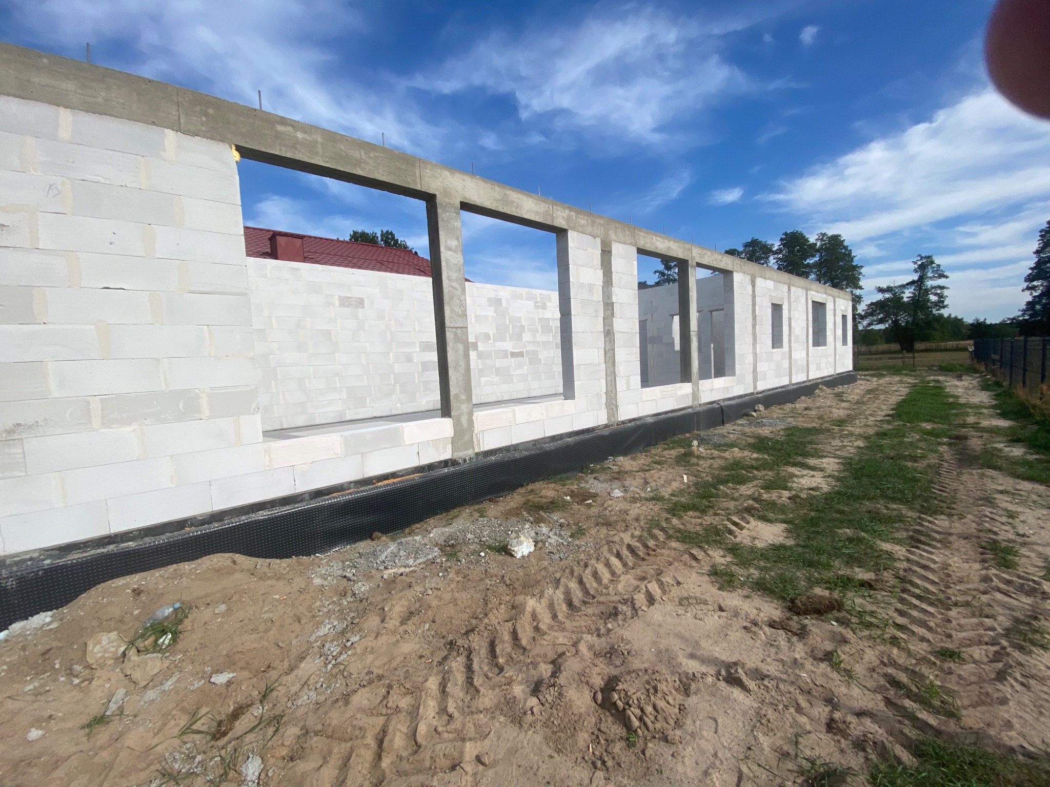 Budowa domu z betonu komórkowego: ściany w trakcie murowania z widocznymi otworami okiennymi i częściowo wylanym wieńcem, z zewnątrz zabezpieczone folią fundamentową, na tle błękitnego nieba.