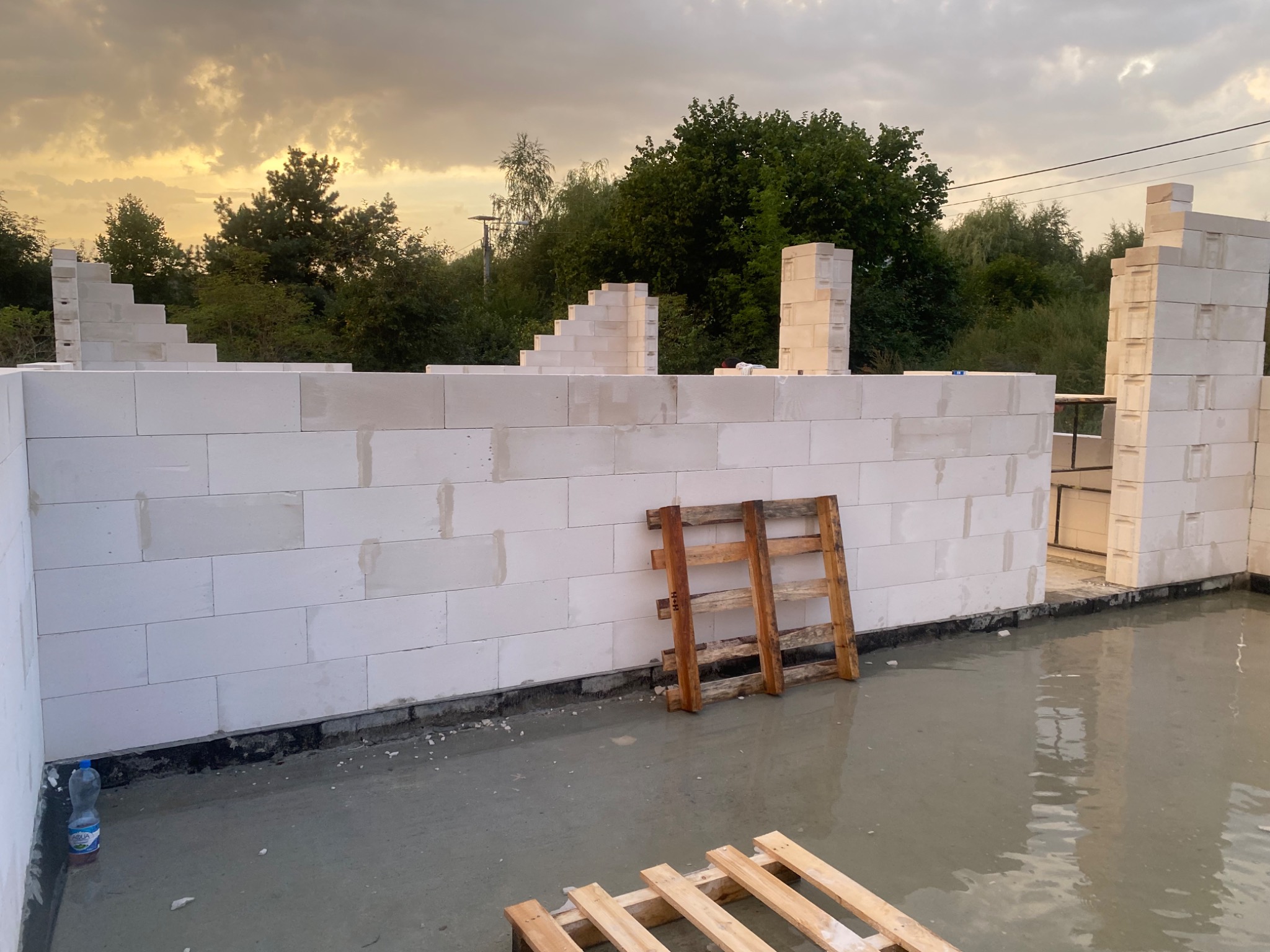 Wznoszenie ścian z bloczków betonu komórkowego na budowie domu w Lipówkach, widoczne palety drewniane i wylany beton na podłodze, pochmurne niebo w tle.