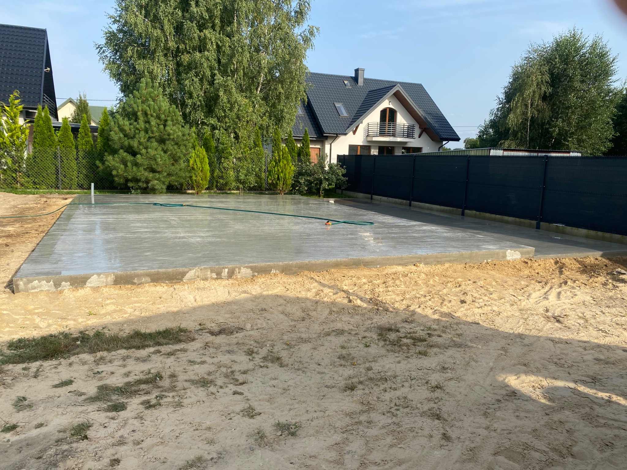 Świeżo wylany, mokry fundament pod budowę domu jednorodzinnego, z widocznym wężem ogrodowym na jego powierzchni, otoczony piaszczystym terenem i ogrodzeniem panelowym.