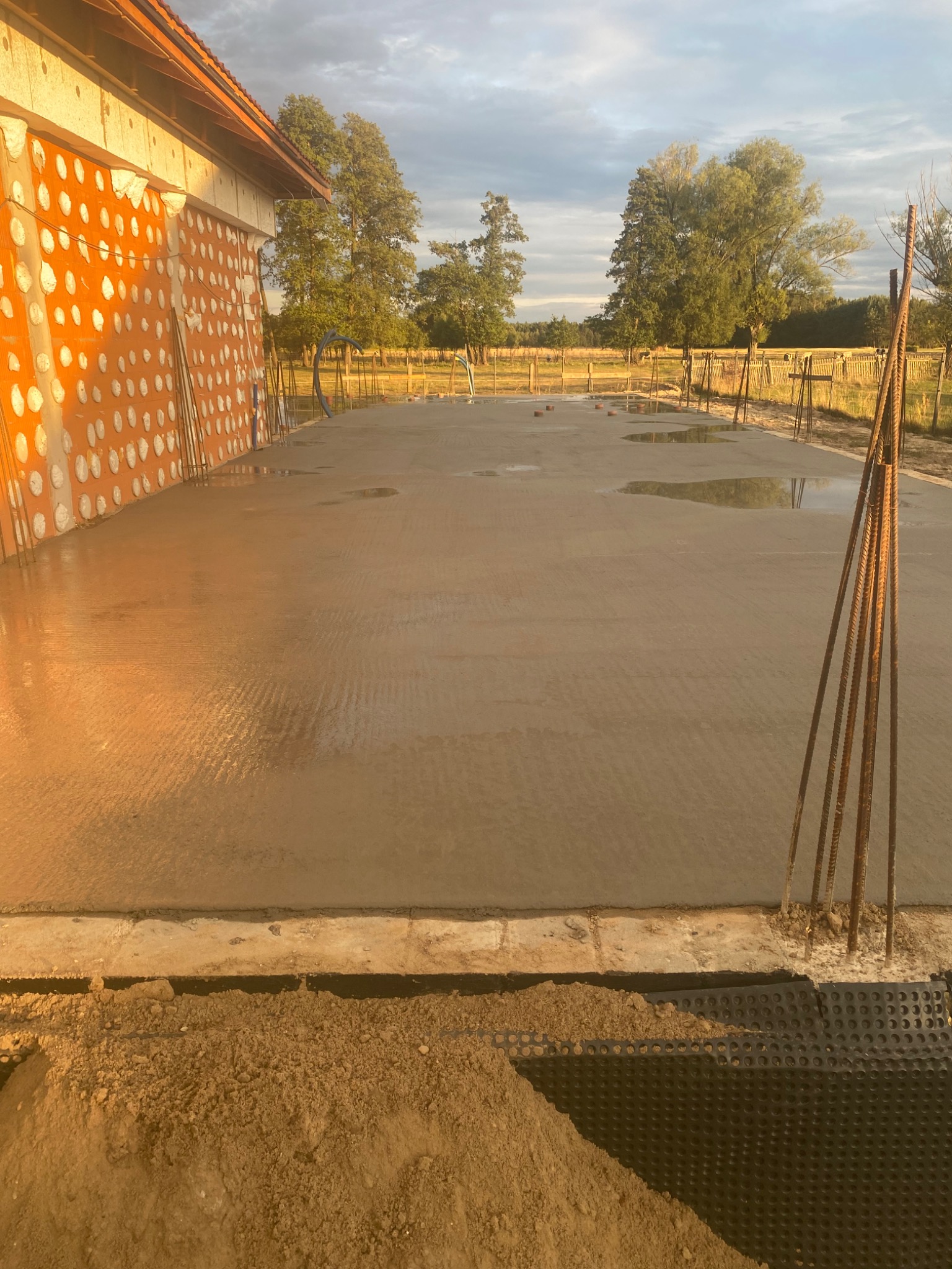 Wylany, świeży betonowy fundament pod budynek z pustaków ceramicznych, widoczne wystające pręty zbrojeniowe i izolacja pionowa.