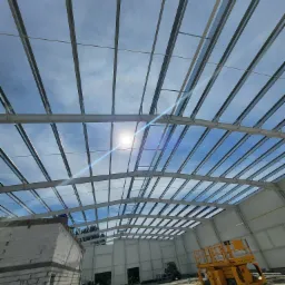 Wnętrze budowanej hali z widoczną konstrukcją stalową dachu, żółtą platformą podnośnikową i fragmentem murowanej ściany z bloczków.