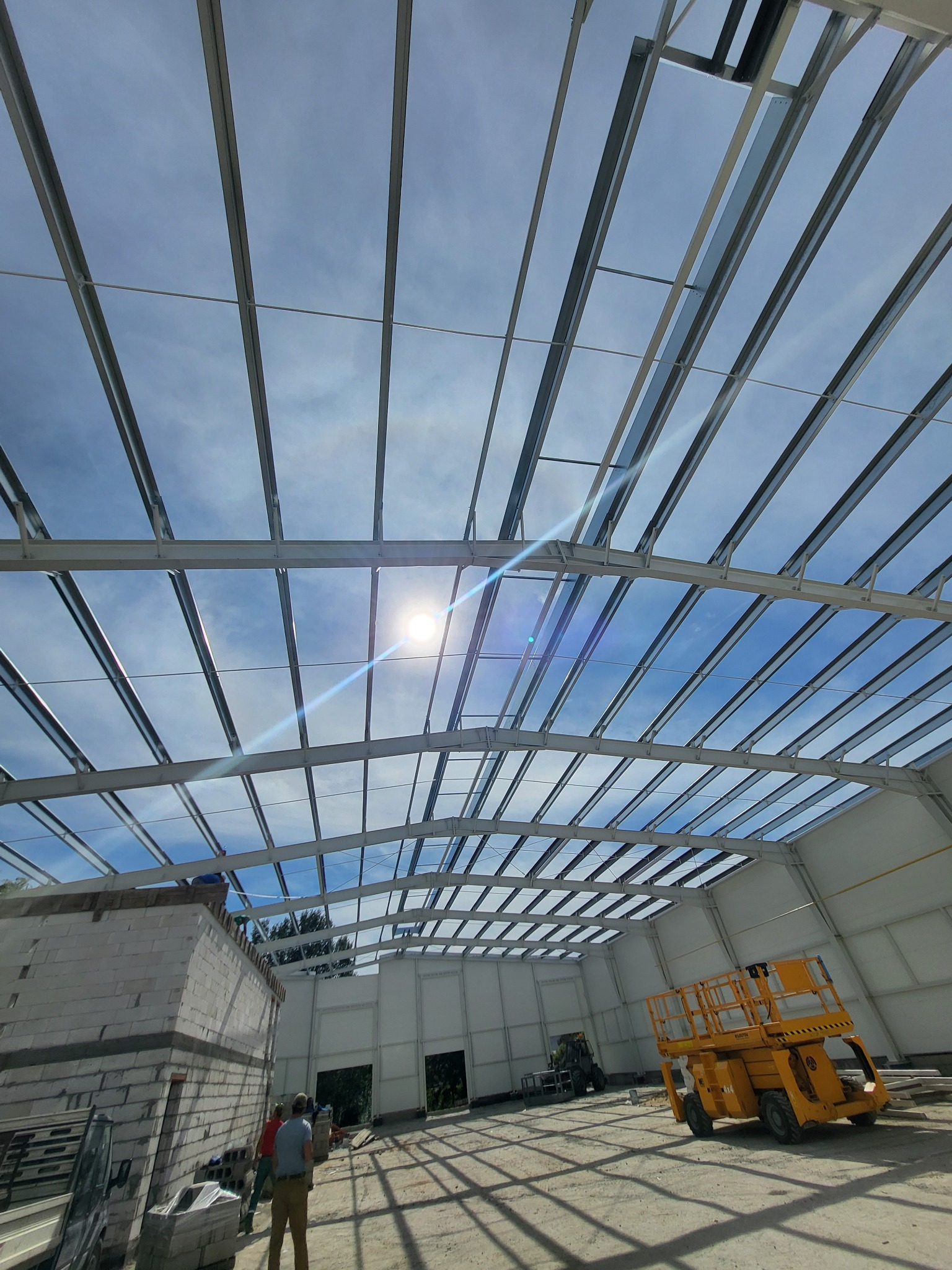 Wnętrze budowanej hali z widoczną konstrukcją stalową dachu, żółtą platformą podnośnikową i fragmentem murowanej ściany z bloczków.