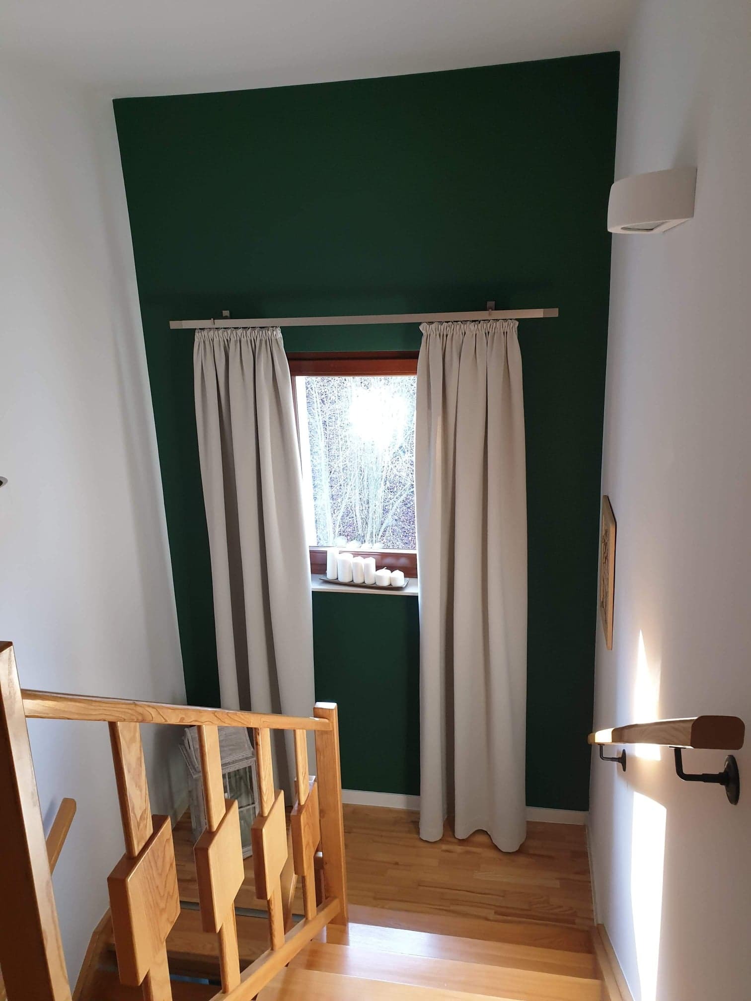 Schody z drewnianą balustradą prowadzą w górę, obok okna z beżowymi zasłonami na tle ściany pomalowanej na butelkową zieleń.