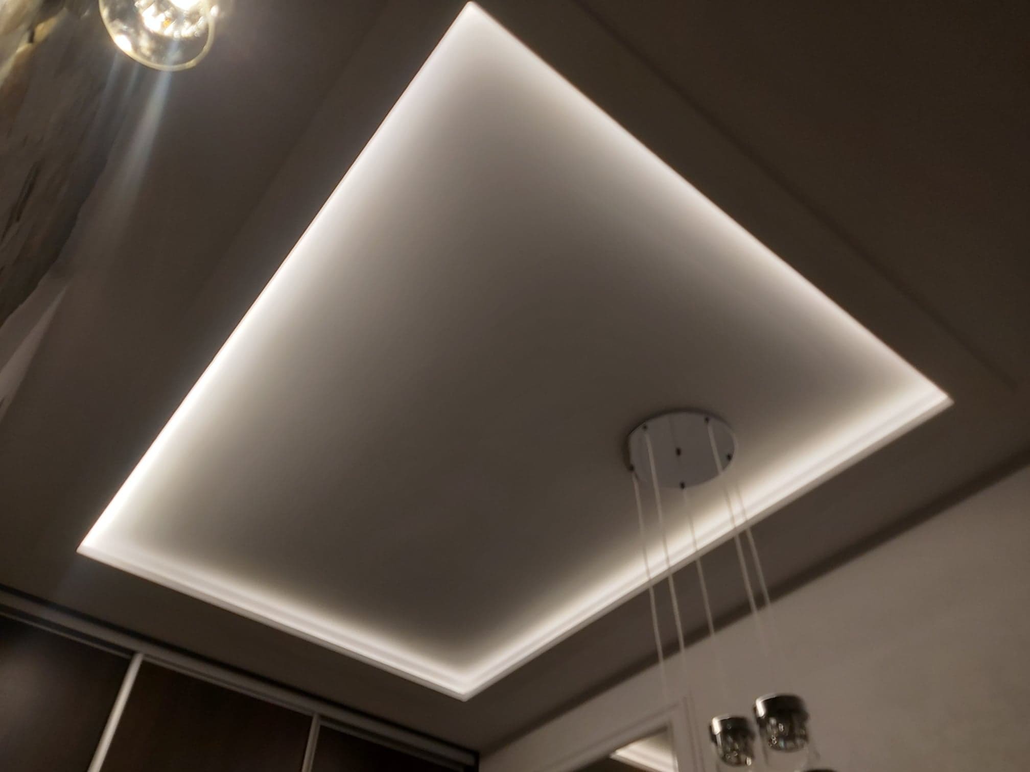 Podwieszany sufit z oświetleniem LED w kształcie prostokąta, widoczne elementy dekoracyjne i fragment lampy wiszącej.