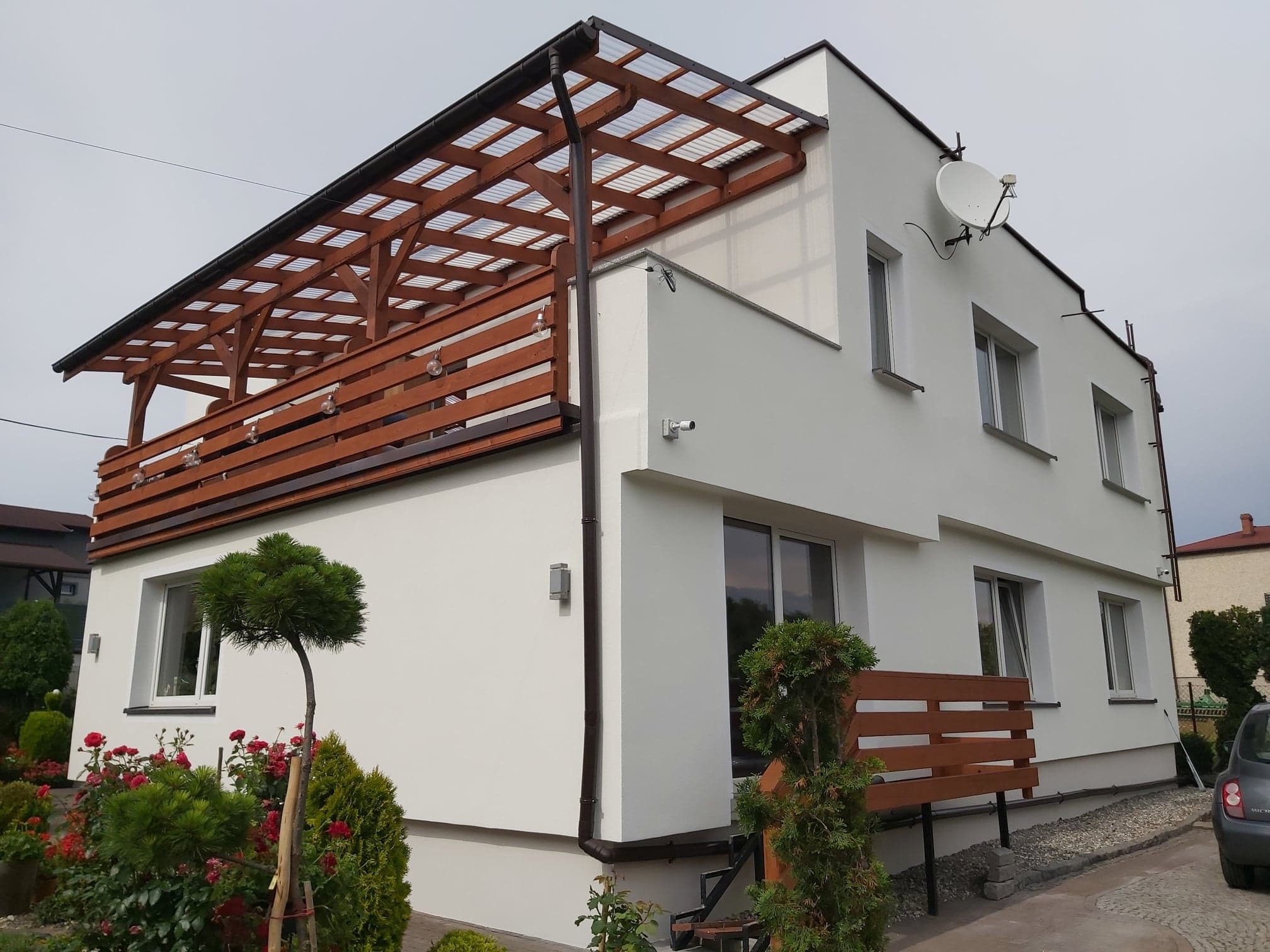 Biały dom jednorodzinny z drewnianą pergolą nad balkonem, ocieplony styropianem, z zamontowaną anteną satelitarną i monitoringiem.