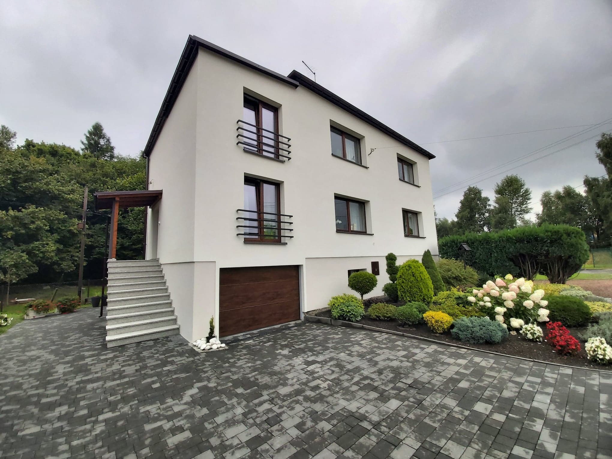 Nowoczesny, dwukondygnacyjny dom z białymi ścianami, brązową stolarką okienną i bramą garażową; widoczny ozdobny ogród z kwitnącymi hortensjami i wybrukowany podjazd z szarej kostki.