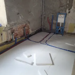 Wnętrze łazienki w trakcie remontu z widoczną instalacją ogrzewania podłogowego, rurami wodnymi i kanalizacyjnymi oraz stelażem podtynkowym WC, ściany w stanie surowym, widoczny styropian...