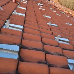 Montaż aluminiowych elementów konstrukcyjnych na pomarańczowej dachówce ceramicznej, przygotowanie do instalacji paneli fotowoltaicznych.