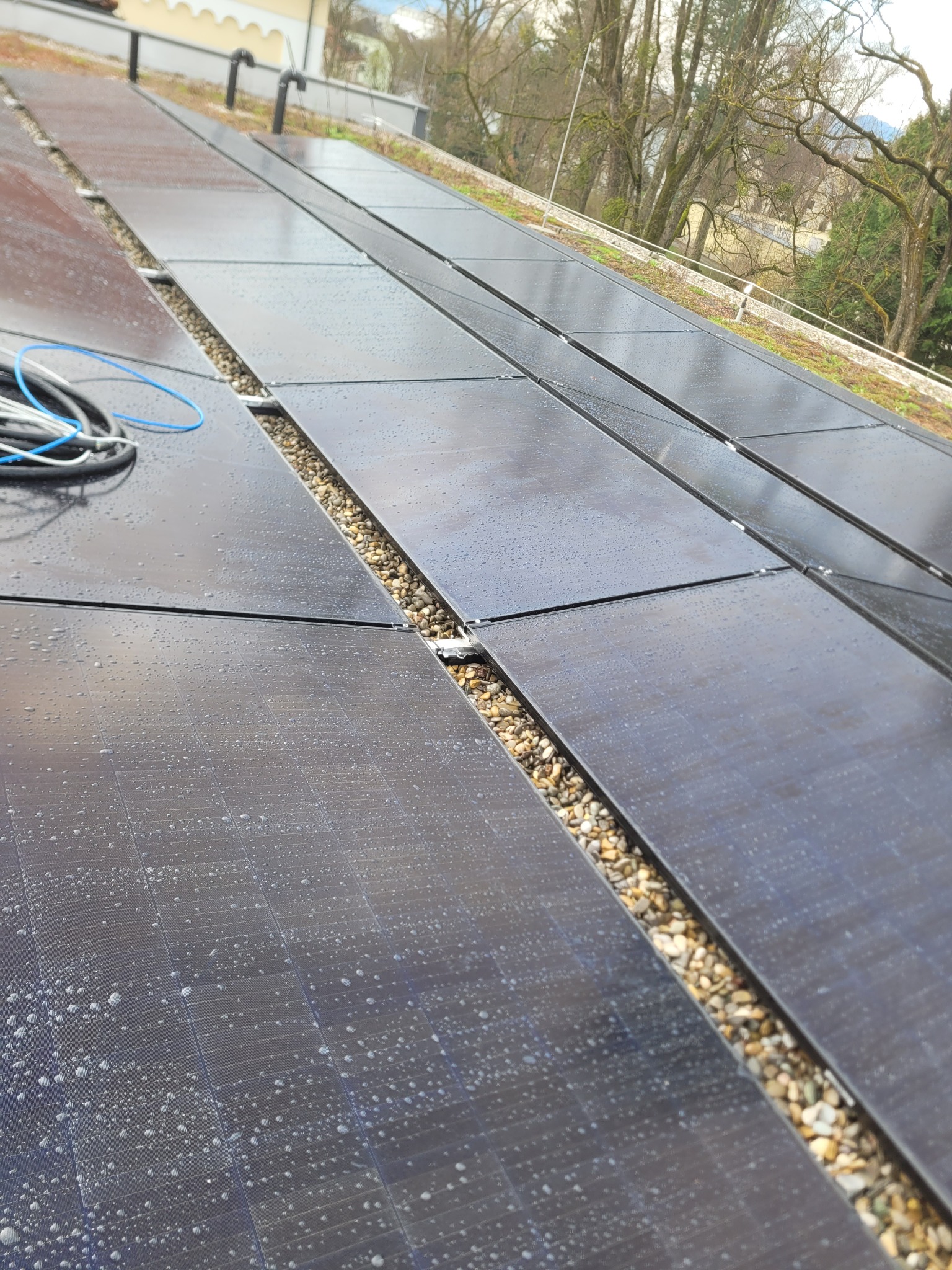 Panel słoneczny na dachu pokryty kroplami wody, widoczne kamyki między panelami i zwój kabli.