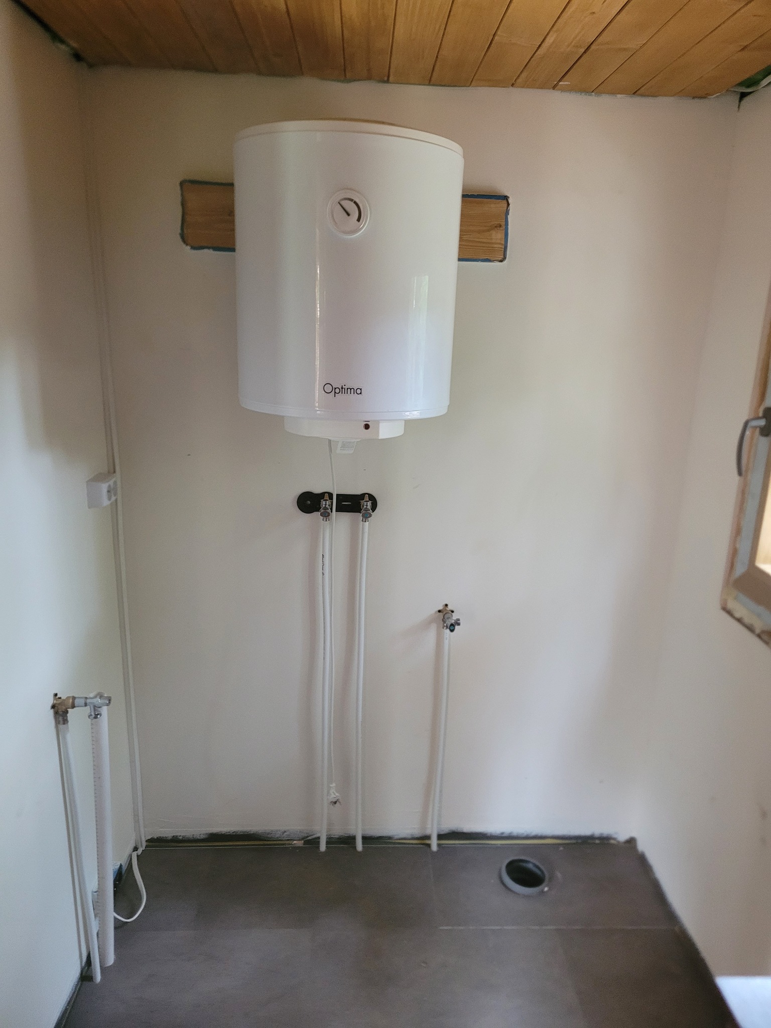 Bojler marki Optima zamontowany na ścianie z widocznymi rurami doprowadzającymi wodę, drewnianym sufitem i oknem w tle.