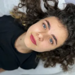 Kobieta z ciemnymi kręconymi włosami i świeżo wystylizowanymi brwiami, leżąca na białym podłożu, skierowana twarzą do góry, z widocznym makijażem ust w kolorze matowej czerwieni.