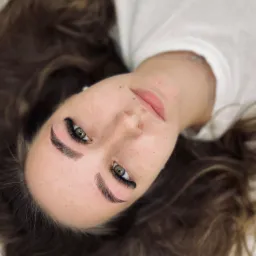 Portret kobiety z zielonymi oczami i świeżo zrobionymi, gęstymi sztucznymi rzęsami w stylu 'kocie oko', leżącej na białym podłożu.