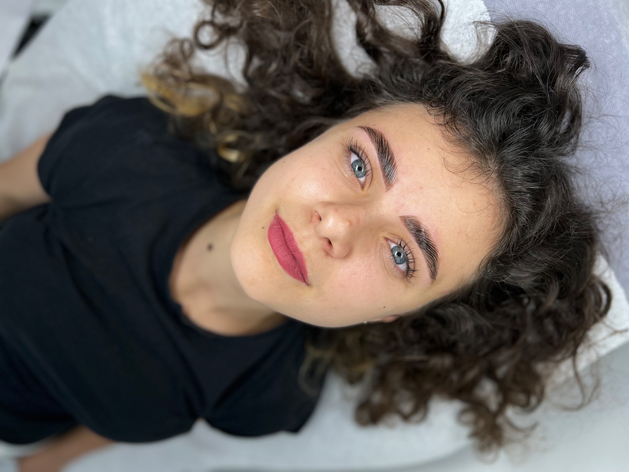 Kobieta z ciemnymi kręconymi włosami i świeżo wystylizowanymi brwiami, leżąca na białym podłożu, skierowana twarzą do góry, z widocznym makijażem ust w kolorze matowej czerwieni.