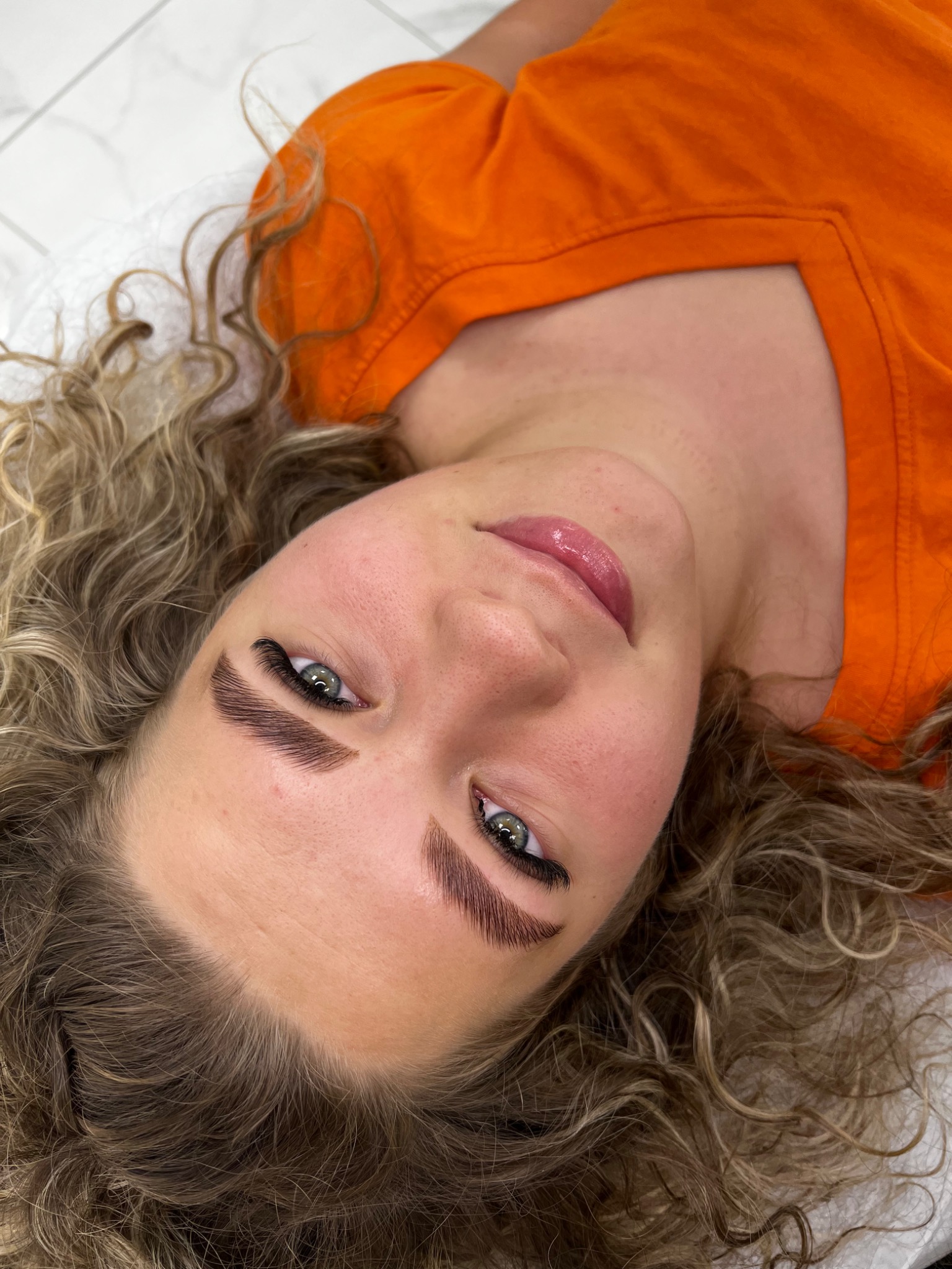 Kobieta z blond lokami leżąca na białym podłożu, prezentująca świeżo wystylizowane brwi i rzęsy.