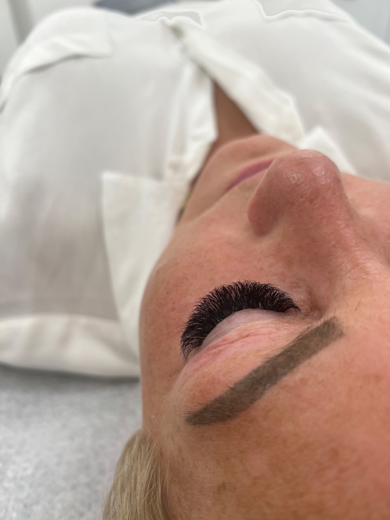 Zbliżenie na oko klientki po zabiegu przedłużania rzęs metodą objętościową, widoczna pełna linia gęstych, czarnych rzęs, lekko podkreślone brwi i fragment białej koszuli w tle.