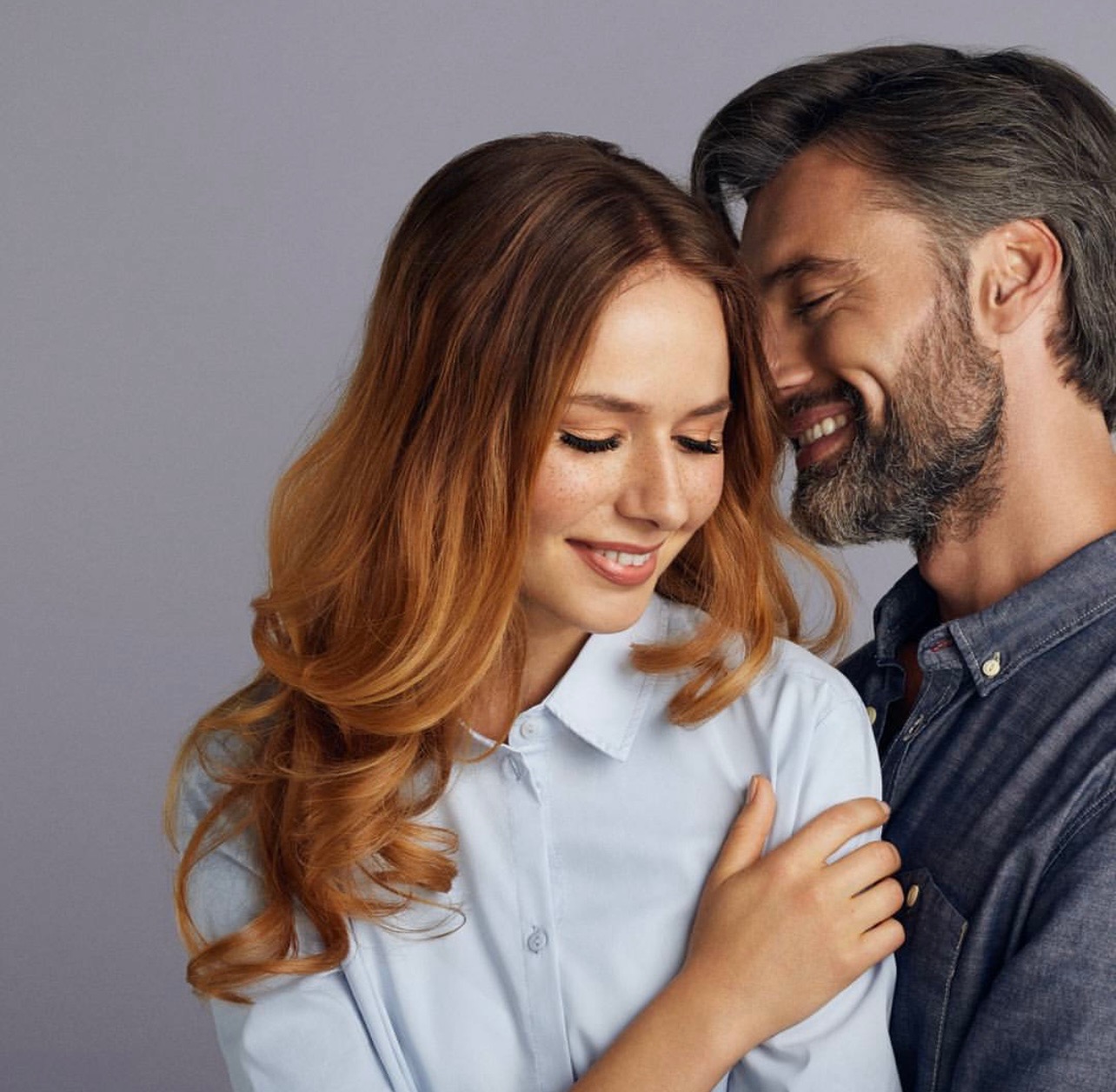 Uśmiechnięta kobieta z rudymi włosami i piegami, obejmowana przez uśmiechniętego mężczyznę z siwą brodą, na jednolitym szarym tle.