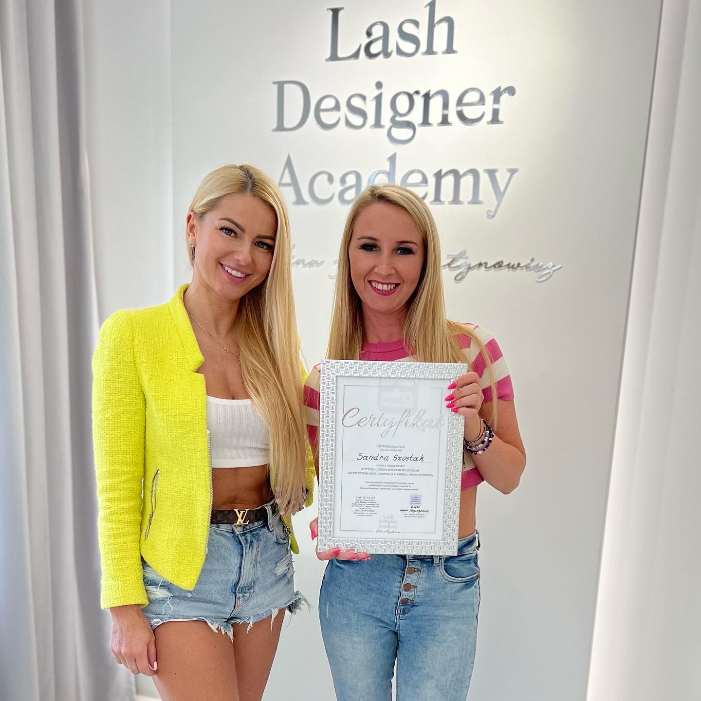 Dwie uśmiechnięte blondynki pozują z certyfikatem Lash Designer Academy na tle logotypu akademii. Jedna w neonowej marynarce i jeansowych szortach, druga w różowo-białej bluzce i jeansach.