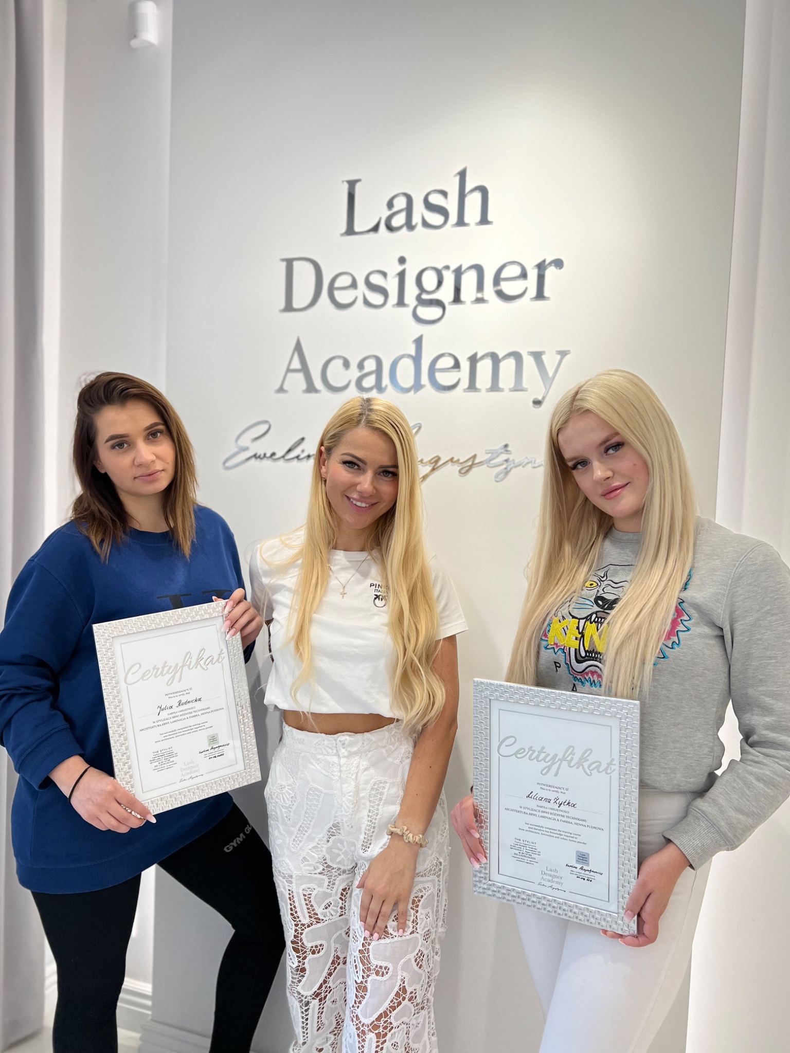 Trzy kobiety pozują z certyfikatami ukończenia kursu Lash Designer Academy na tle ściany z logo akademii.