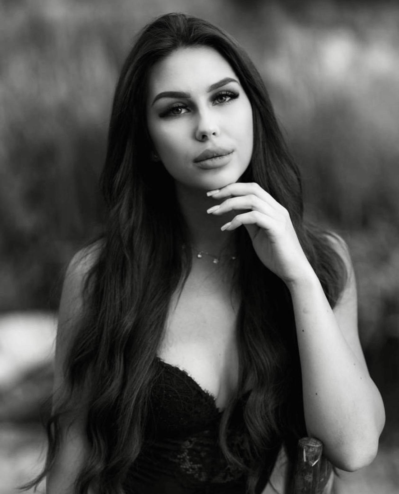 Portret kobiety z długimi, falowanymi włosami, delikatnym makijażem i naszyjnikiem, w czarno-białej tonacji, z ręką pod brodą, oparta o drewniany element.