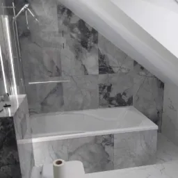 Nowoczesna łazienka na poddaszu z marmurowymi płytkami w odcieniach szarości, biała wanna zabudowana płytkami, toaleta z rolką papieru na pokrywie, kabina prysznicowa ze szklaną ścianką.