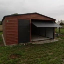 Nowoczesny, brązowy garaż z blachy trapezowej, z czarnymi drzwiami uchylnymi i bocznymi, w pochmurny dzień, na trawiastym terenie.