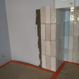 Układanie pionowych, błyszczących płytek w odcieniach beżu i szarości na ścianie w kuchni, widoczne krzyżyki dystansowe, fragment ściany przygotowany pod płytki, zabezpieczona pomarańczową folią...