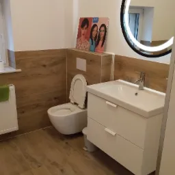 Nowoczesna łazienka we Wrocławiu z drewnopodobnymi płytkami, okrągłym lustrem z podświetleniem LED, białą umywalką z szafką i toaletą.