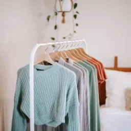 Rząd ubrań w pastelowych kolorach, wiszących na drewnianych wieszakach na białym metalowym stojaku, z rośliną w makramie wiszącą w tle.