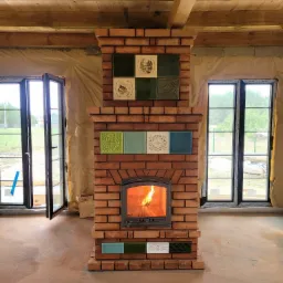 Murowany kominek z czerwonej cegły z dekoracyjnymi kaflami w odcieniach zieleni i błękitu, płomień w palenisku, surowy stan wykończenia wnętrza z drewnianym stropem i folią na ścianach, otwarte okna.