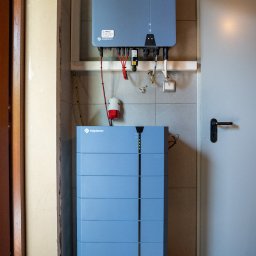 NOVUS ENERGY SPÓŁKA Z OGRANICZONĄ ODPOWIEDZIALNOŚCIĄ - Niebieski magazyn energii Solplanet zamontowany na ścianie z szarymi płytkami, obok białe drzwi. Widoczne kable i elementy instalacji elektrycznej.