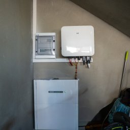NOVUS ENERGY SPÓŁKA Z OGRANICZONĄ ODPOWIEDZIALNOŚCIĄ - Biały magazyn energii GoodWe, inwerter i skrzynka elektryczna zamontowane na szarej ścianie. Widoczne okablowanie i elementy instalacji. U dołu fragment roweru i akcesoriów.