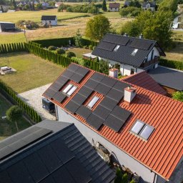 NOVUS ENERGY SPÓŁKA Z OGRANICZONĄ ODPOWIEDZIALNOŚCIĄ - Widok z góry na domy z panelami słonecznymi na dachach. Panele na dachu z czerwoną dachówką. W tle zabudowa jednorodzinna i tereny zielone.