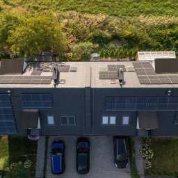 NOVUS ENERGY SPÓŁKA Z OGRANICZONĄ ODPOWIEDZIALNOŚCIĄ - Widok z góry na szereg domów z panelami słonecznymi na dachu i elewacji, z trzema samochodami zaparkowanymi przed budynkami. W tle zieleń.