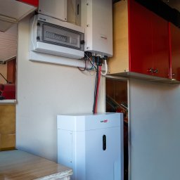 NOVUS ENERGY SPÓŁKA Z OGRANICZONĄ ODPOWIEDZIALNOŚCIĄ - Biały magazyn energii SolarEdge w pomieszczeniu z szafkami, obok niego widoczny inwerter i rozdzielnia elektryczna. Kable prowadzą do urządzeń.