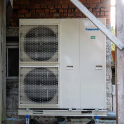 NOVUS ENERGY SPÓŁKA Z OGRANICZONĄ ODPOWIEDZIALNOŚCIĄ - Zewnętrzna jednostka pompy ciepła Panasonic R32, zamontowana przy ceglanym budynku, z widocznymi dwoma wentylatorami i fragmentem drewnianej konstrukcji.