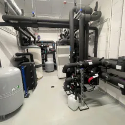 Węzeł cieplny w kotłowni: rury w izolacji, zbiorniki wyrównawcze Reflex, pompy obiegowe Grundfos, białe ściany z bloków, szara posadzka, instalacje pod sufitem.
