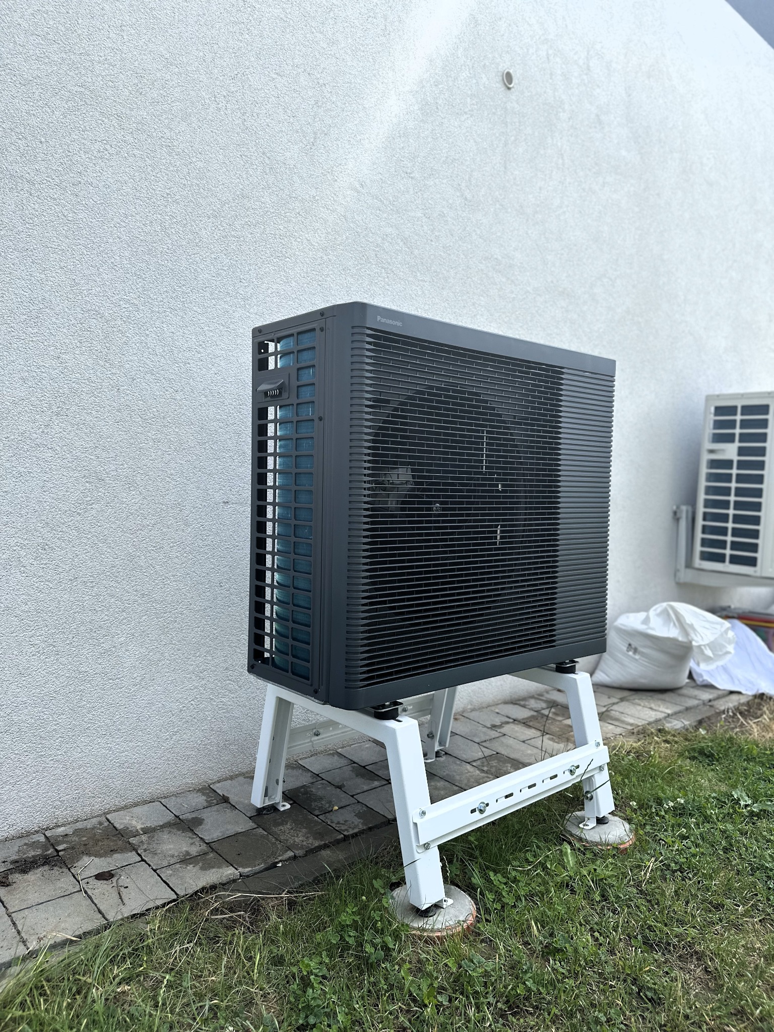 Zewnętrzna jednostka pompy ciepła Panasonic zamontowana na białym stelażu przy ścianie budynku, z widoczną drugą jednostką klimatyzacji w tle i workami ochronnymi.