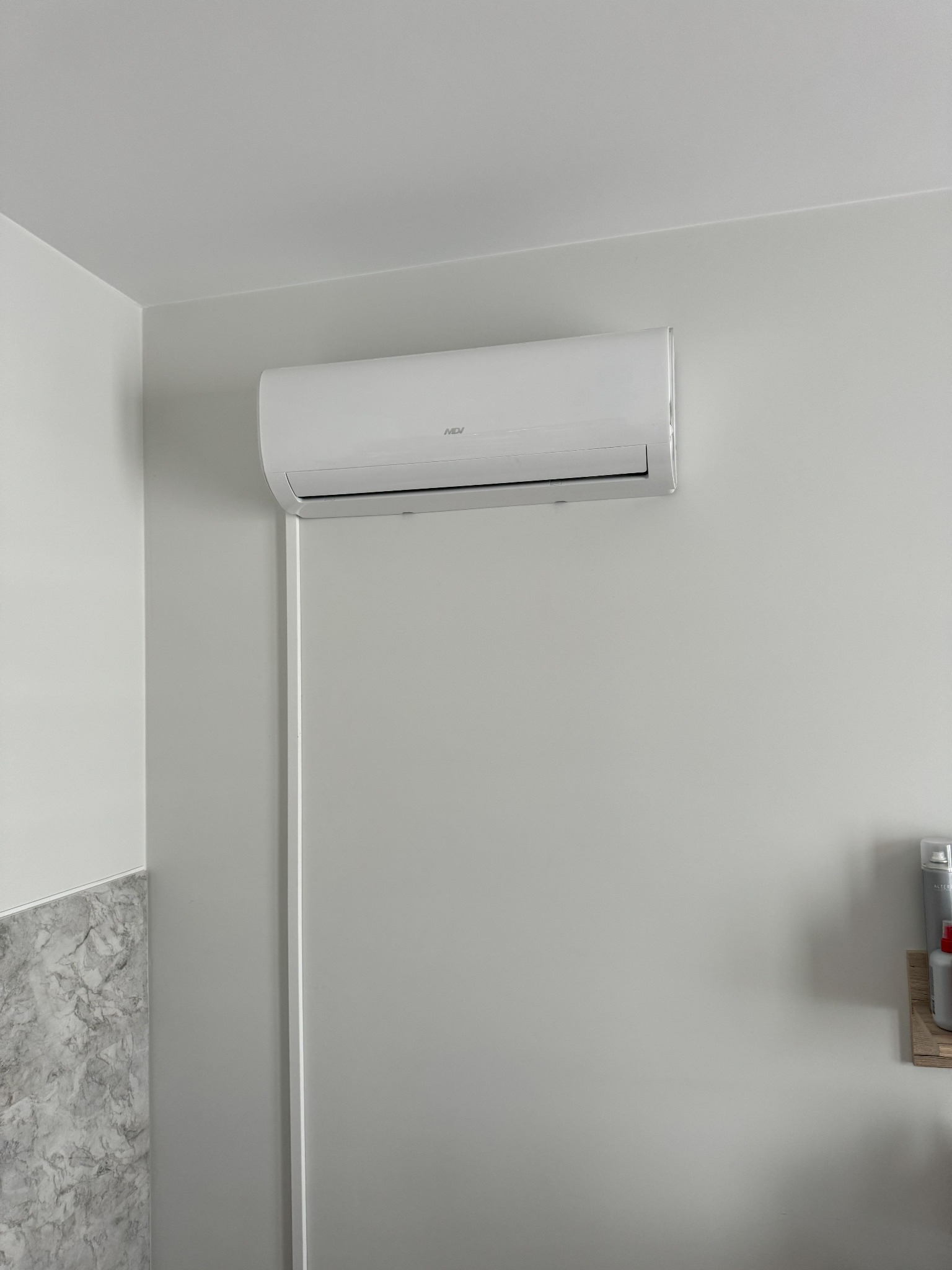 Biała klimatyzacja ścienna marki MDJ zamontowana w jasnym pomieszczeniu, widoczna część dekoracyjnej ściany z efektem marmuru po lewej stronie i kosmetyki na półce po prawej.