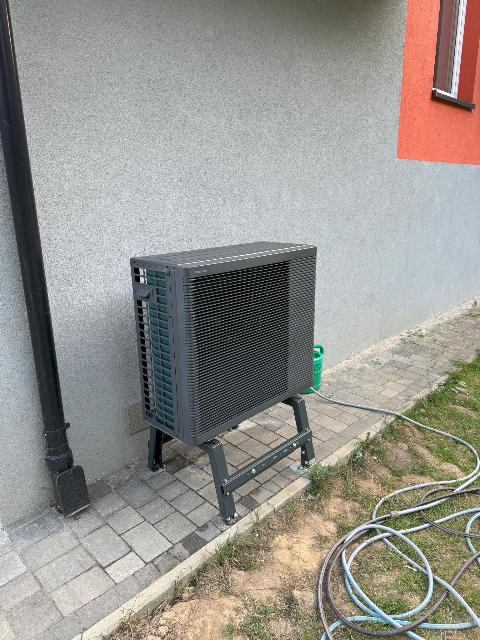 Szara jednostka zewnętrzna pompy ciepła Panasonic, umieszczona na metalowym stojaku, przy szarej ścianie budynku z fragmentem pomarańczowej elewacji i widoczną rurą spustową, obok chodnika z kostki...