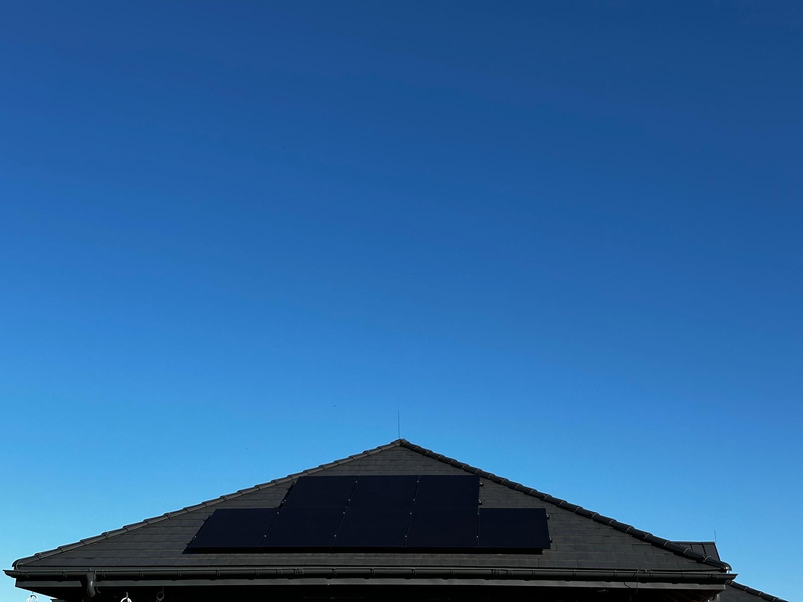 Ciemne panele słoneczne na dachu domu z ciemnej dachówki, na tle intensywnie błękitnego nieba i widocznym piorunochronem.