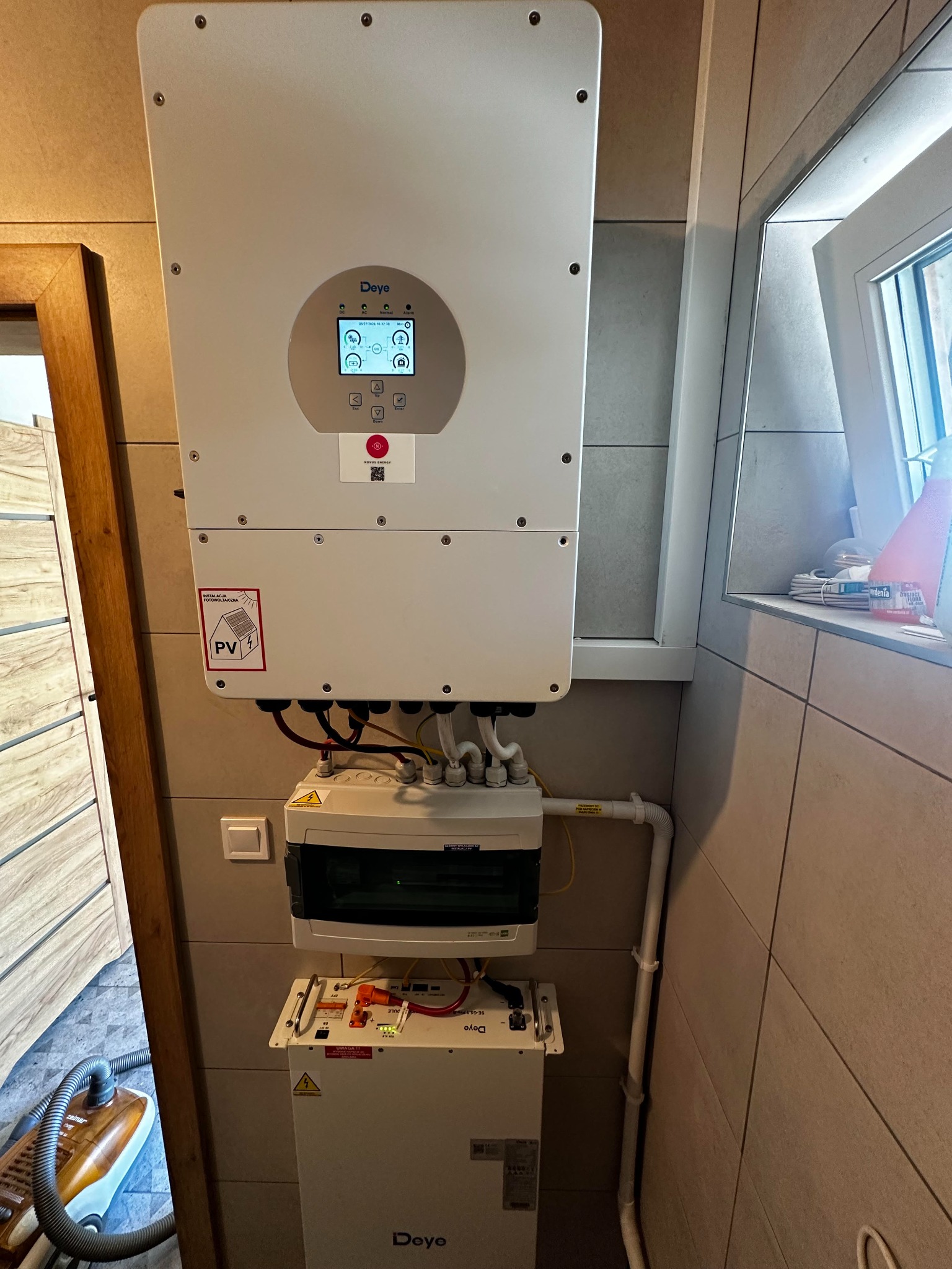 Instalacja domowa: Falownik marki Deye z wyświetlaczem, skrzynka elektryczna i magazyn energii Deye, zamontowane na kafelkowanej ścianie, obok okna i drewnianej framugi drzwi.