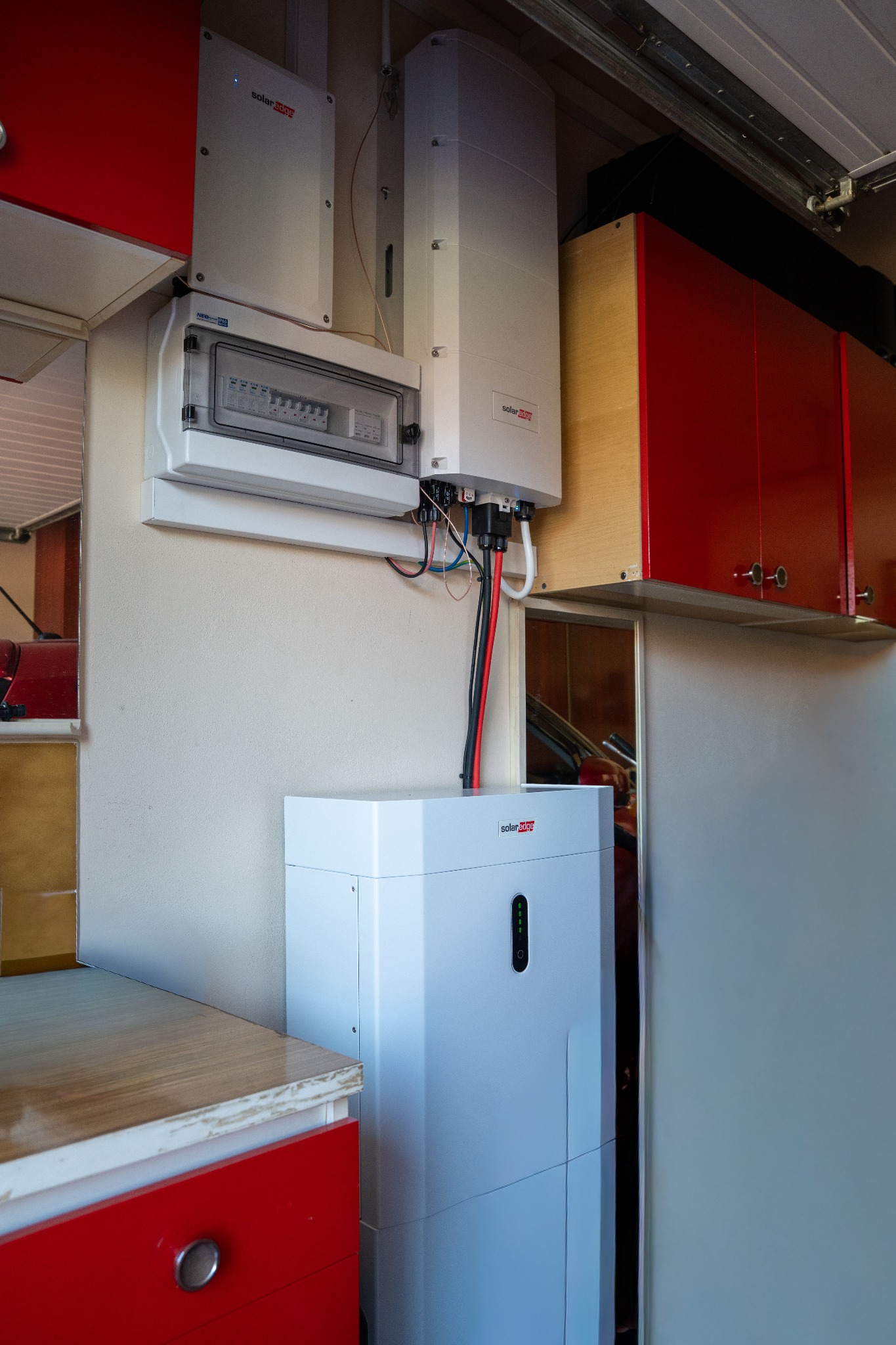 Domowa instalacja: biały magazyn energii SolarEdge obok skrzynki elektrycznej i inwertera, z czerwonymi szafkami w tle. Układ w pomieszczeniu.