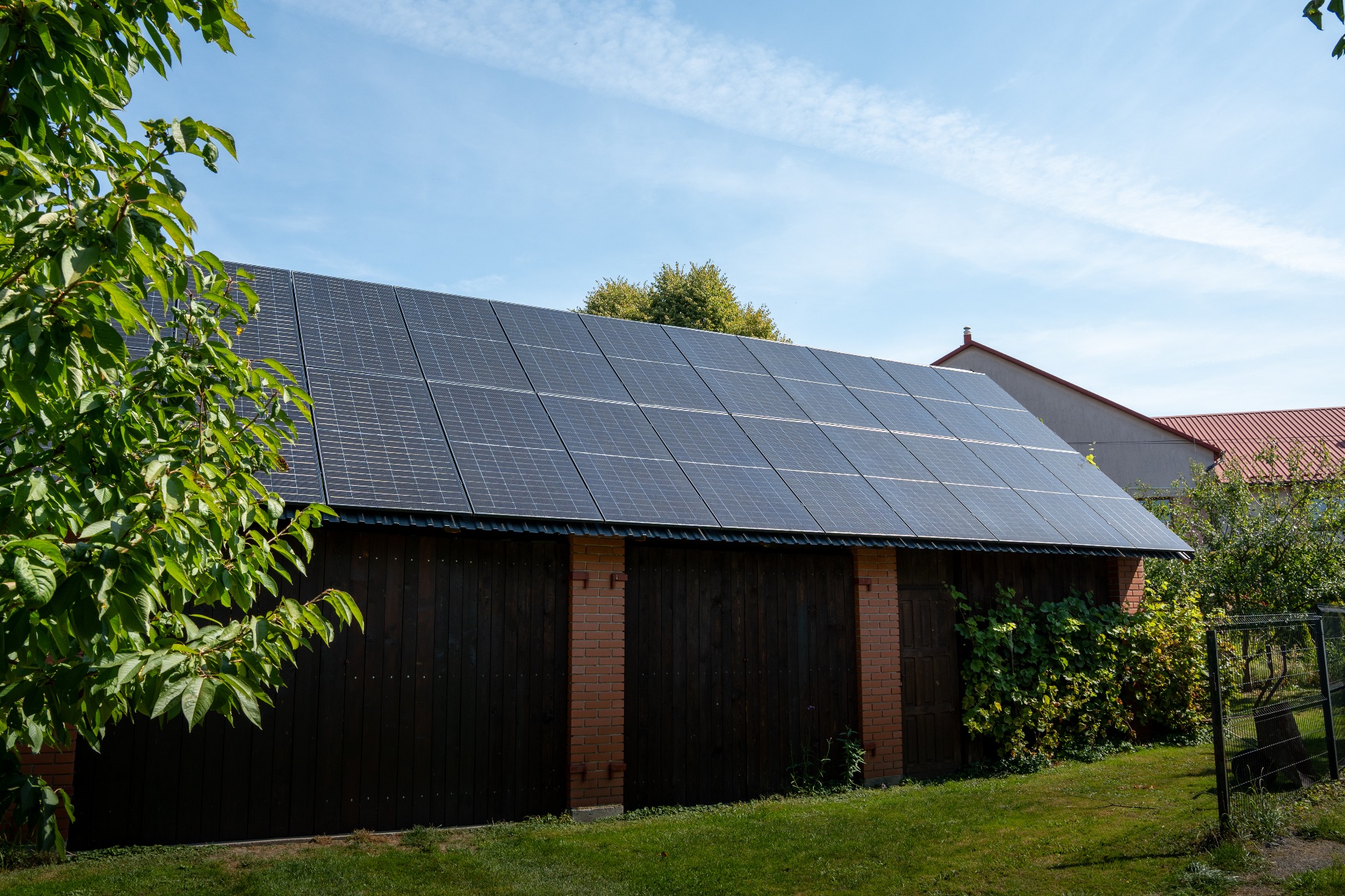Instalacja paneli słonecznych na dachu budynku gospodarczego z ciemnego drewna, widok z poziomu trawnika w słoneczny dzień. W tle budynki mieszkalne.