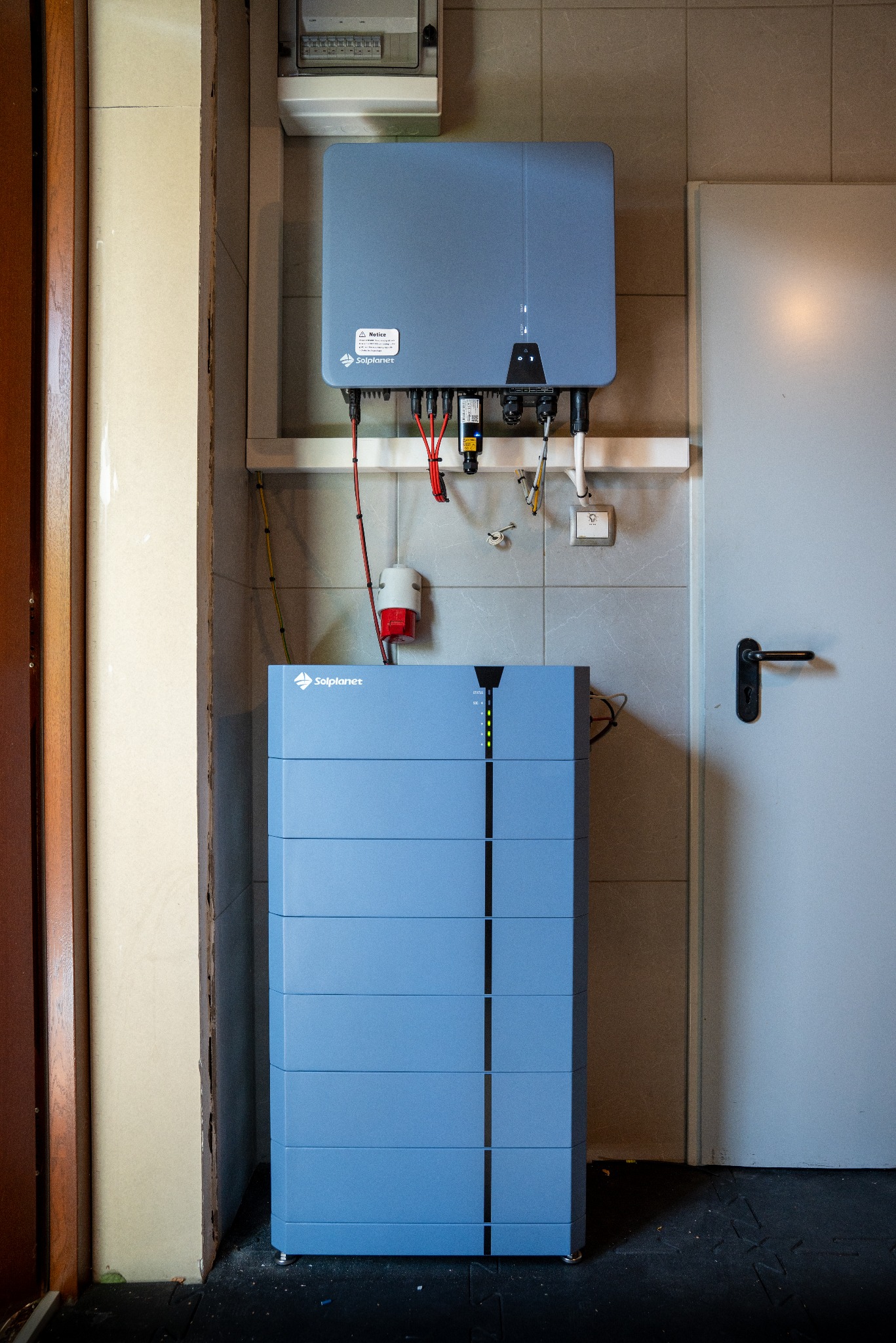 Niebieski magazyn energii Solplanet zamontowany na ścianie z szarymi płytkami, obok białe drzwi. Widoczne kable i elementy instalacji elektrycznej.