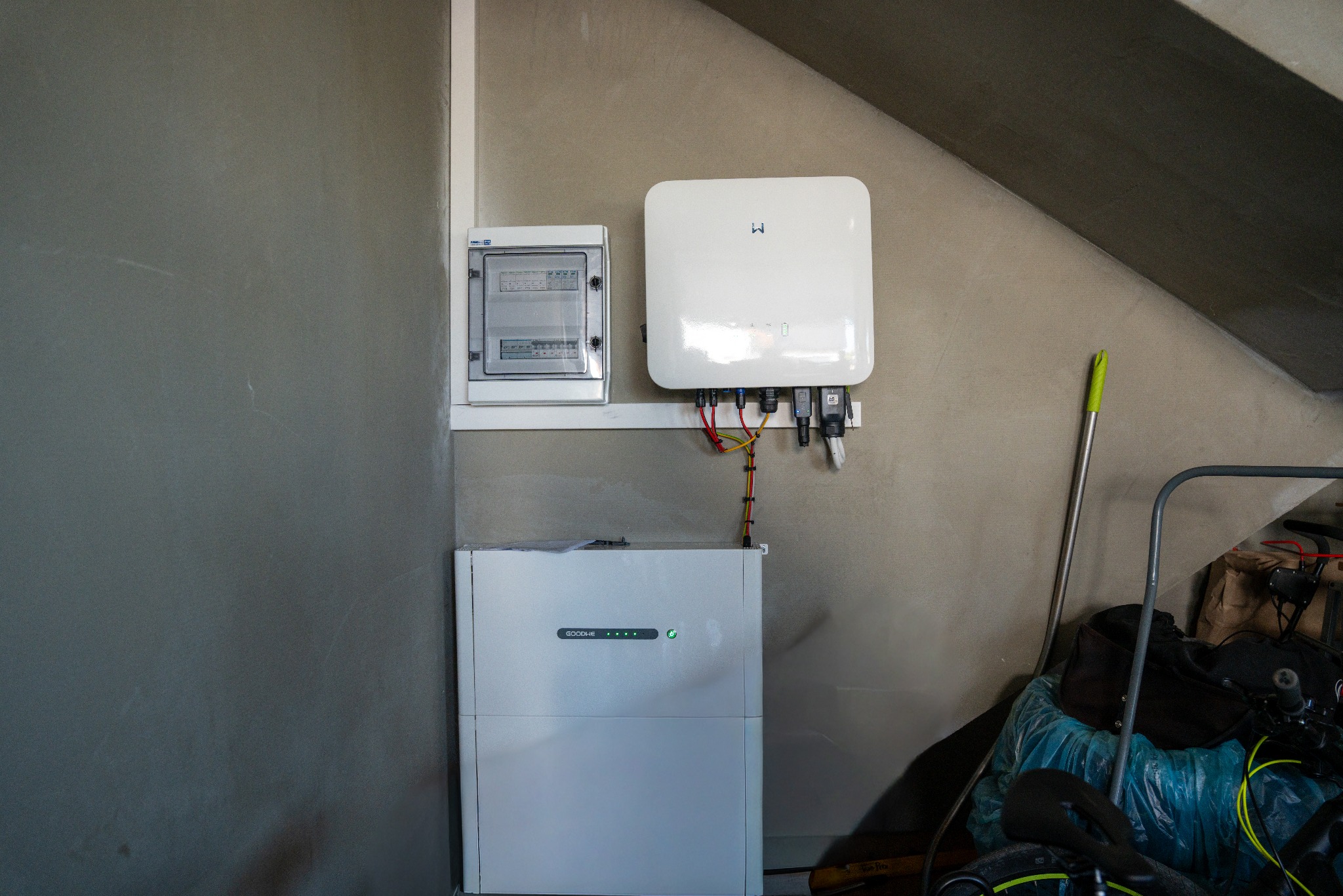 Biały magazyn energii GoodWe, inwerter i skrzynka elektryczna zamontowane na szarej ścianie. Widoczne okablowanie i elementy instalacji. U dołu fragment roweru i akcesoriów.