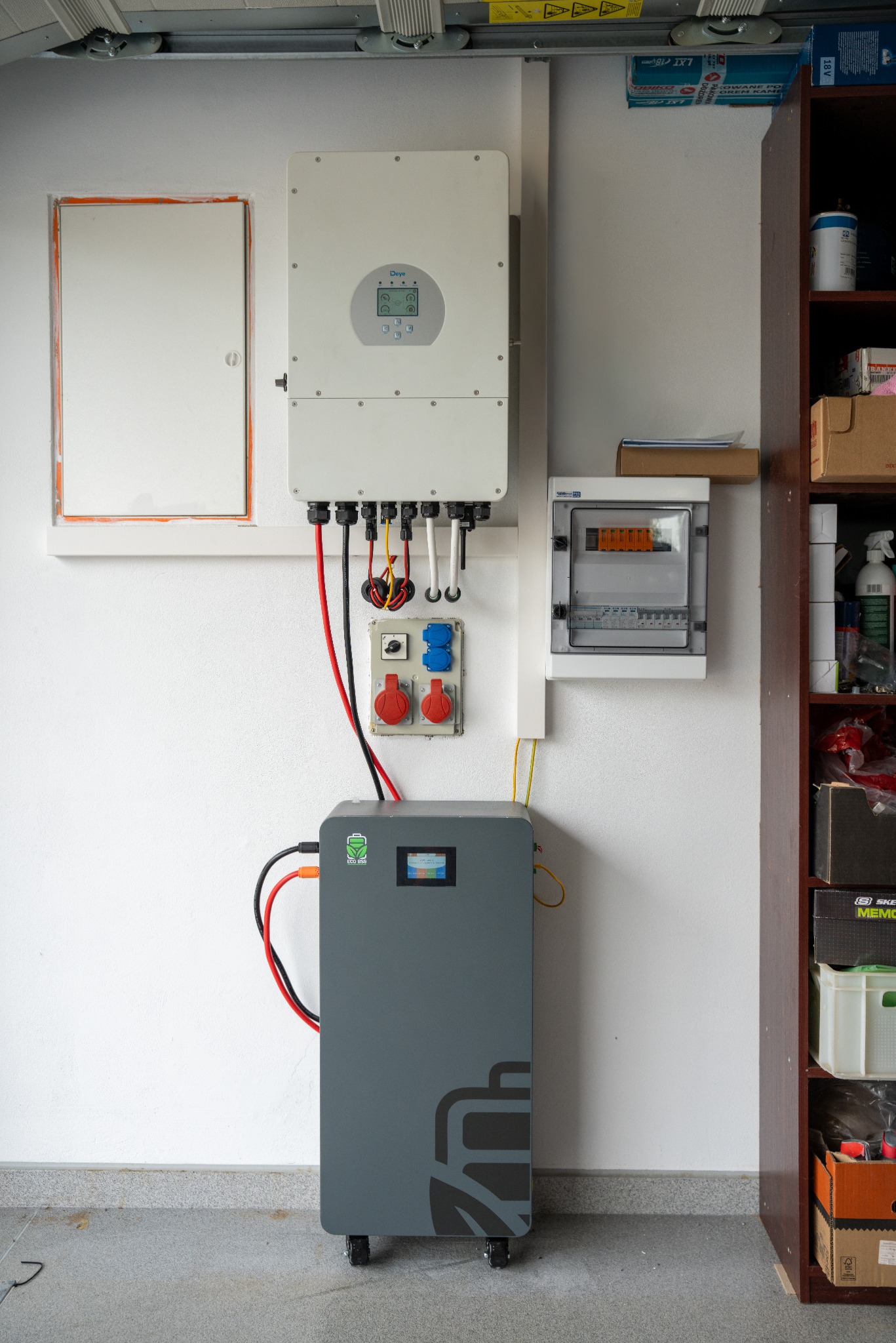Szary magazyn energii na kółkach z podłączonym inwerterem na ścianie, obok skrzynka elektryczna i gniazda siłowe. Elementy instalacji w garażu.