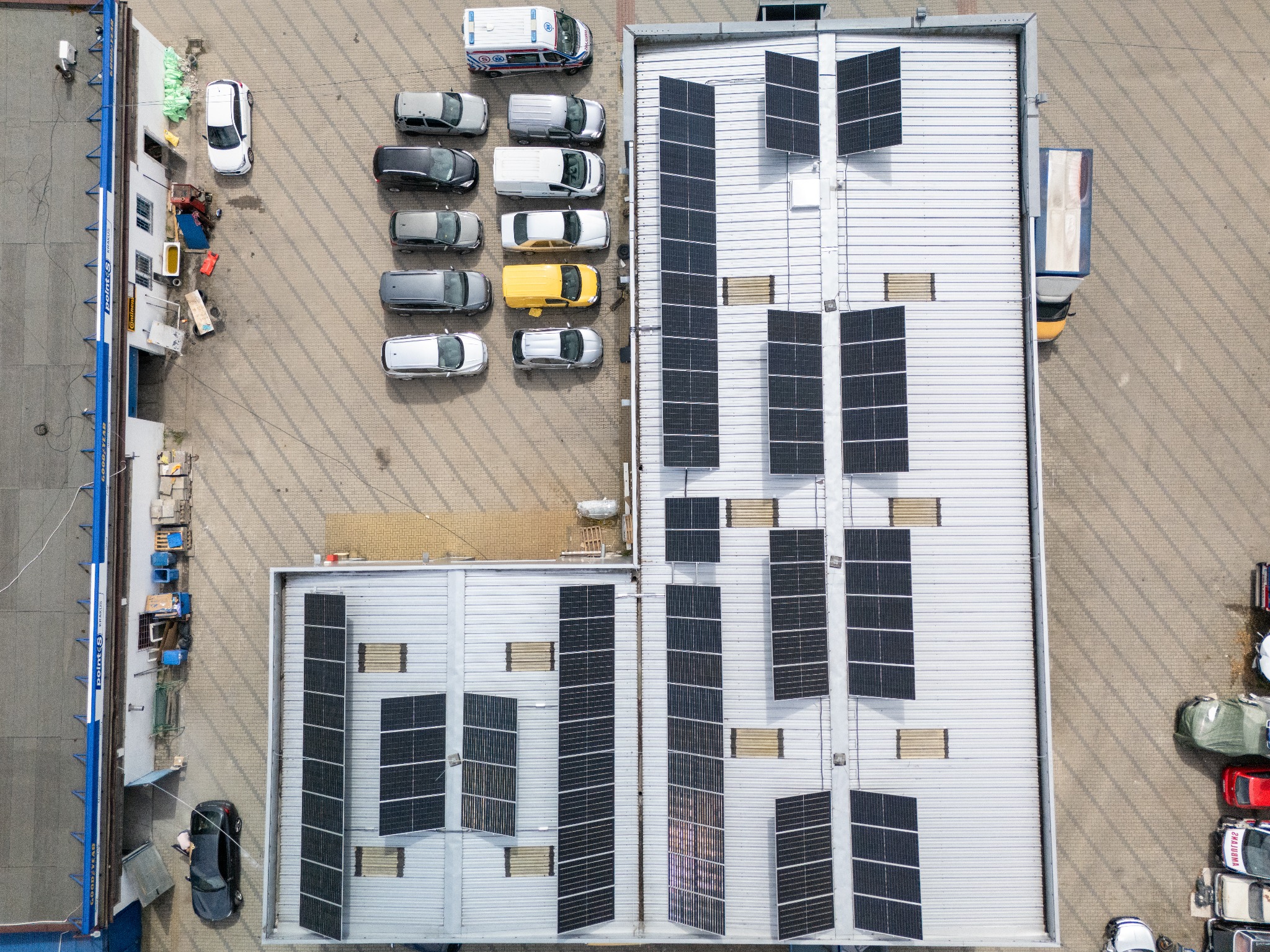 Widok z góry na dach budynku z panelami słonecznymi, obok zaparkowane samochody, w tym ambulans. Panele ułożone w rzędach, widoczne detale konstrukcji dachu.