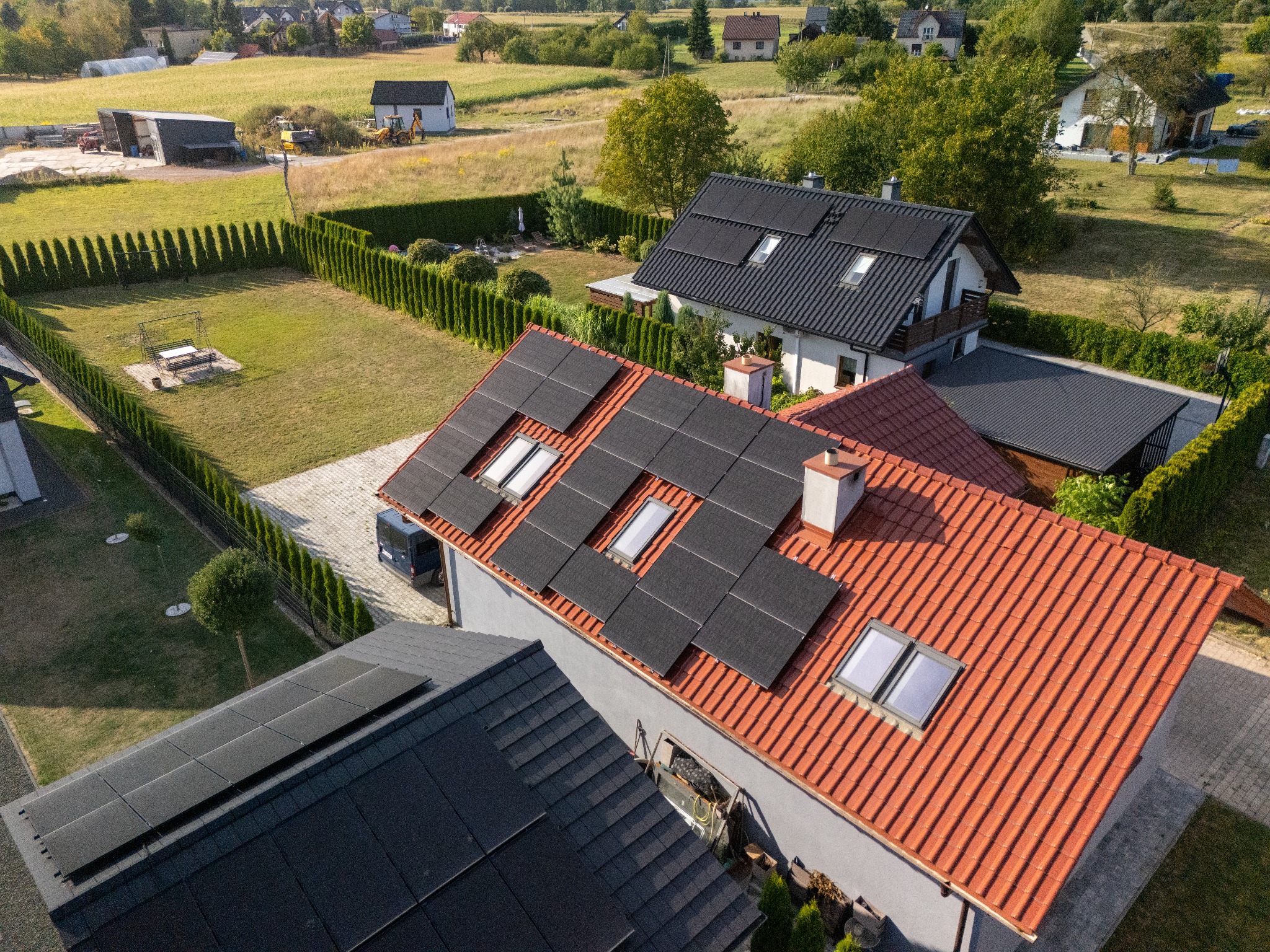 Widok z góry na domy z panelami słonecznymi na dachach. Panele na dachu z czerwoną dachówką. W tle zabudowa jednorodzinna i tereny zielone.