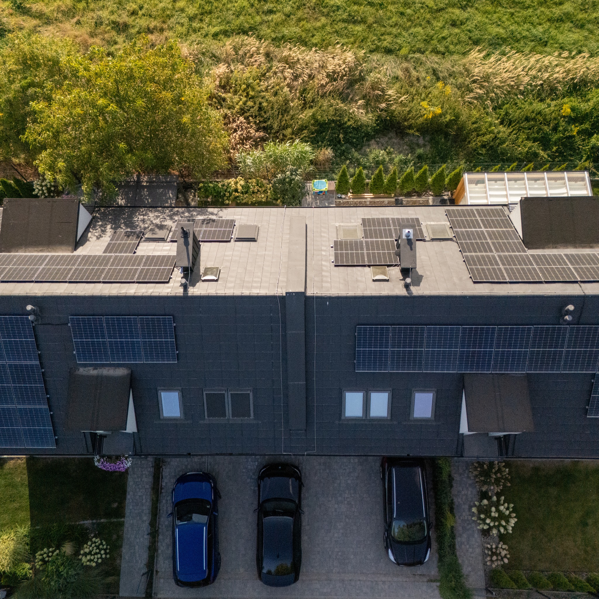 Widok z góry na szereg domów z panelami słonecznymi na dachu i elewacji, z trzema samochodami zaparkowanymi przed budynkami. W tle zieleń.